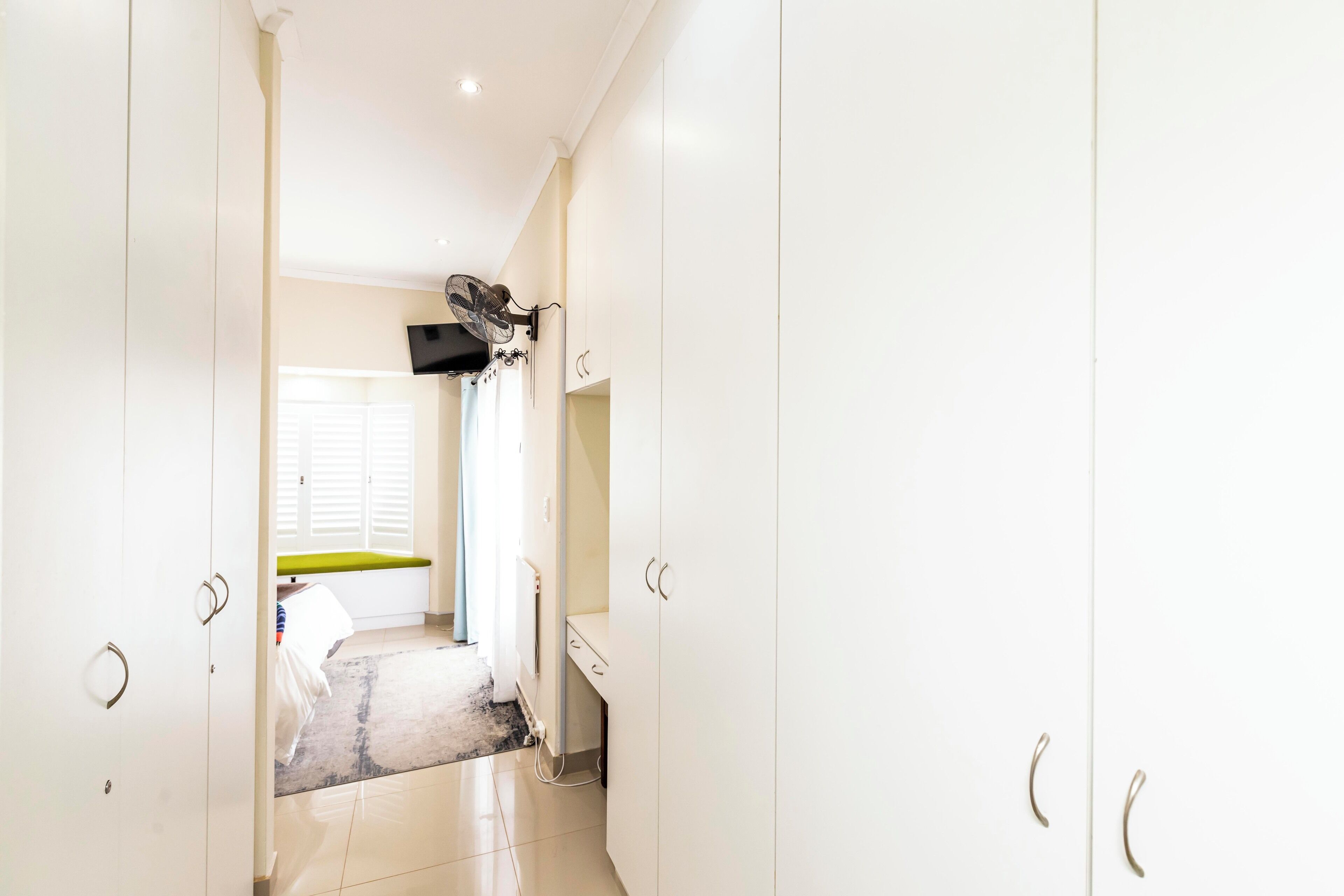 A hallway with white cabinets, a television mounted on the wall, and a window with blinds.