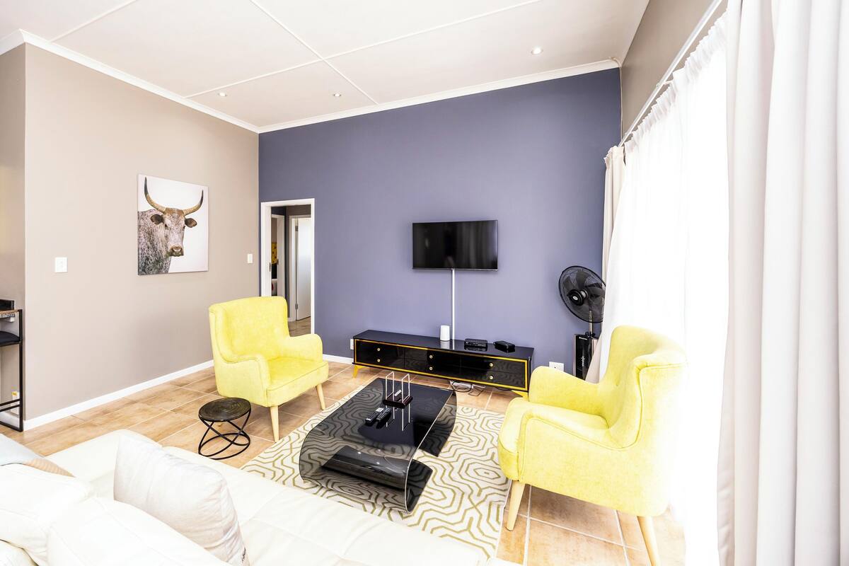 Un salón moderno con un televisor de pantalla plana, sillones amarillos, una mesa de centro negra y un cuadro decorativo de cabeza de toro en la pared.