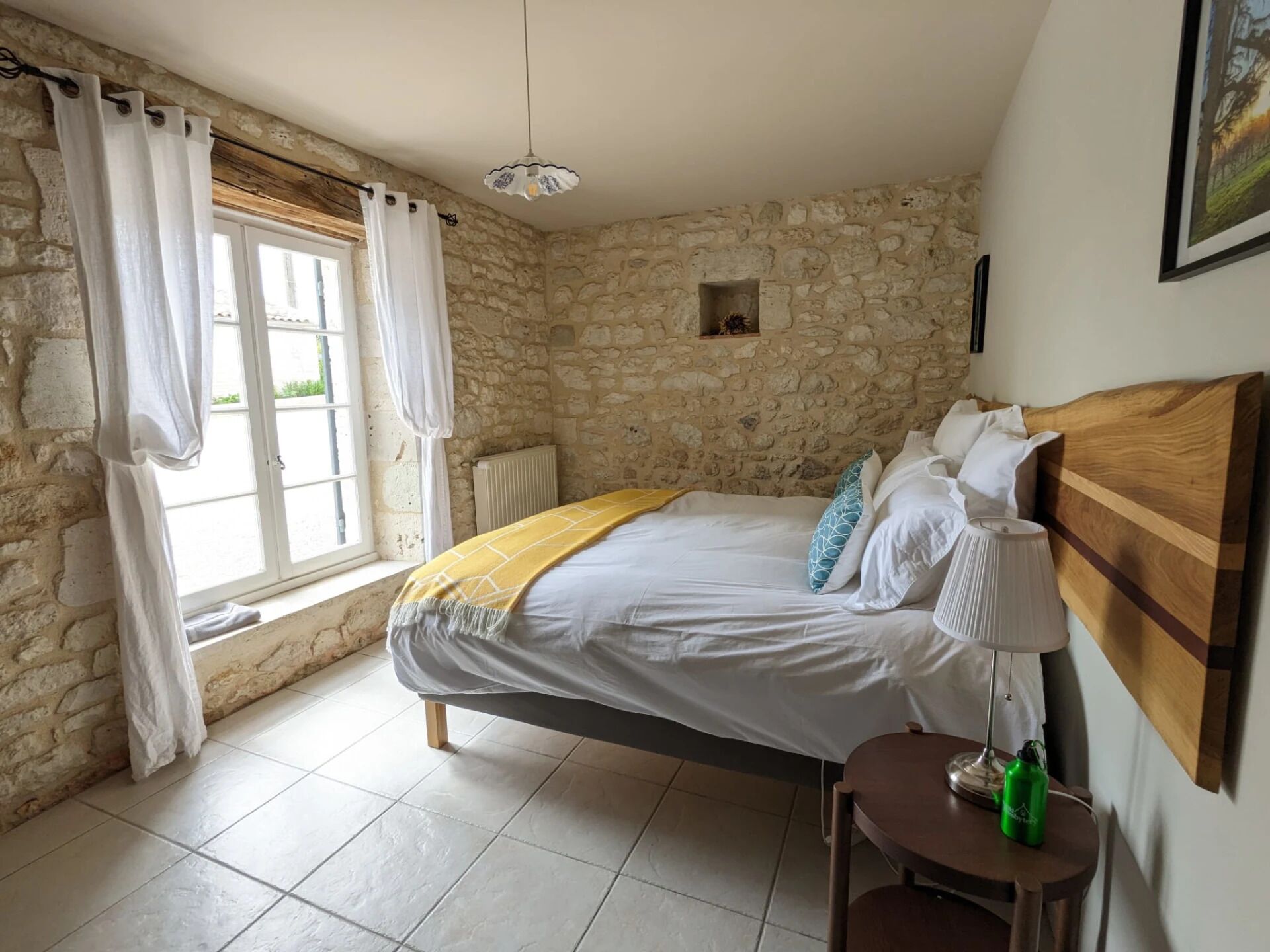 A bedroom with a stone wall, a bed with white bedding, a wooden headboard, a window with white curtains, and a small table with a lamp and a green bottle.