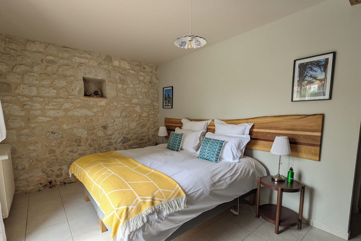 Una camera da letto con parete in pietra, testiera in legno, letto con biancheria bianca e cuscini decorati, comodino con lampada e un quadro appeso al muro.