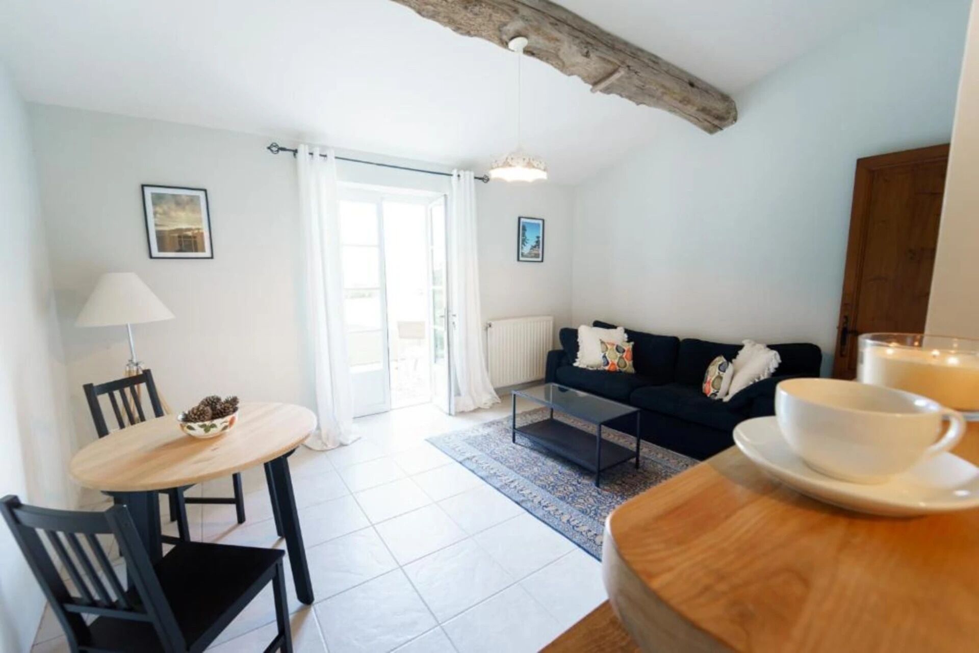 Una sala de estar moderna con un sofá, una mesa de comedor con sillas, una mesa de centro y una lámpara. Hay una ventana con cortinas, un radiador y un cuadro en la pared.