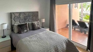 A bedroom with a bed, nightstands, a ceiling light, and a view of an outdoor area with chairs and a palm tree. - Ground floor South facing apartment (Murcia)