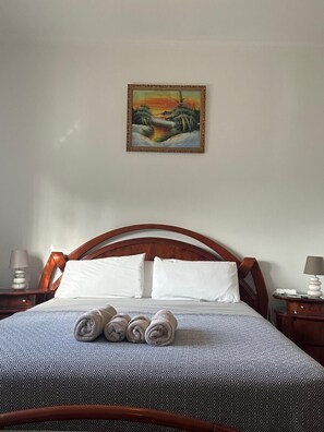 A neatly made bed with a patterned bedspread, two white pillows, and rolled towels at the foot of the bed. A painting of a landscape hangs above the bed.