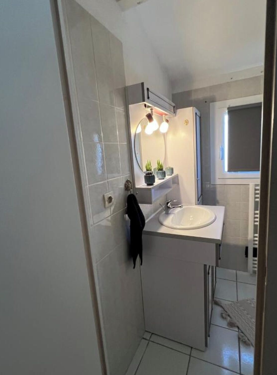 Une salle de bain avec un lavabo blanc, un miroir avec éclairage intégré, un porte-serviettes et une fenêtre.