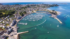 Vista aérea de un pueblo costero con un puerto deportivo, zonas residenciales y un puerto curvo destacado.
