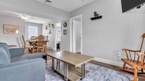 Un salon comprenant un canapé, une table basse, un coin repas avec des chaises, un téléviseur fixé au mur et une horloge.