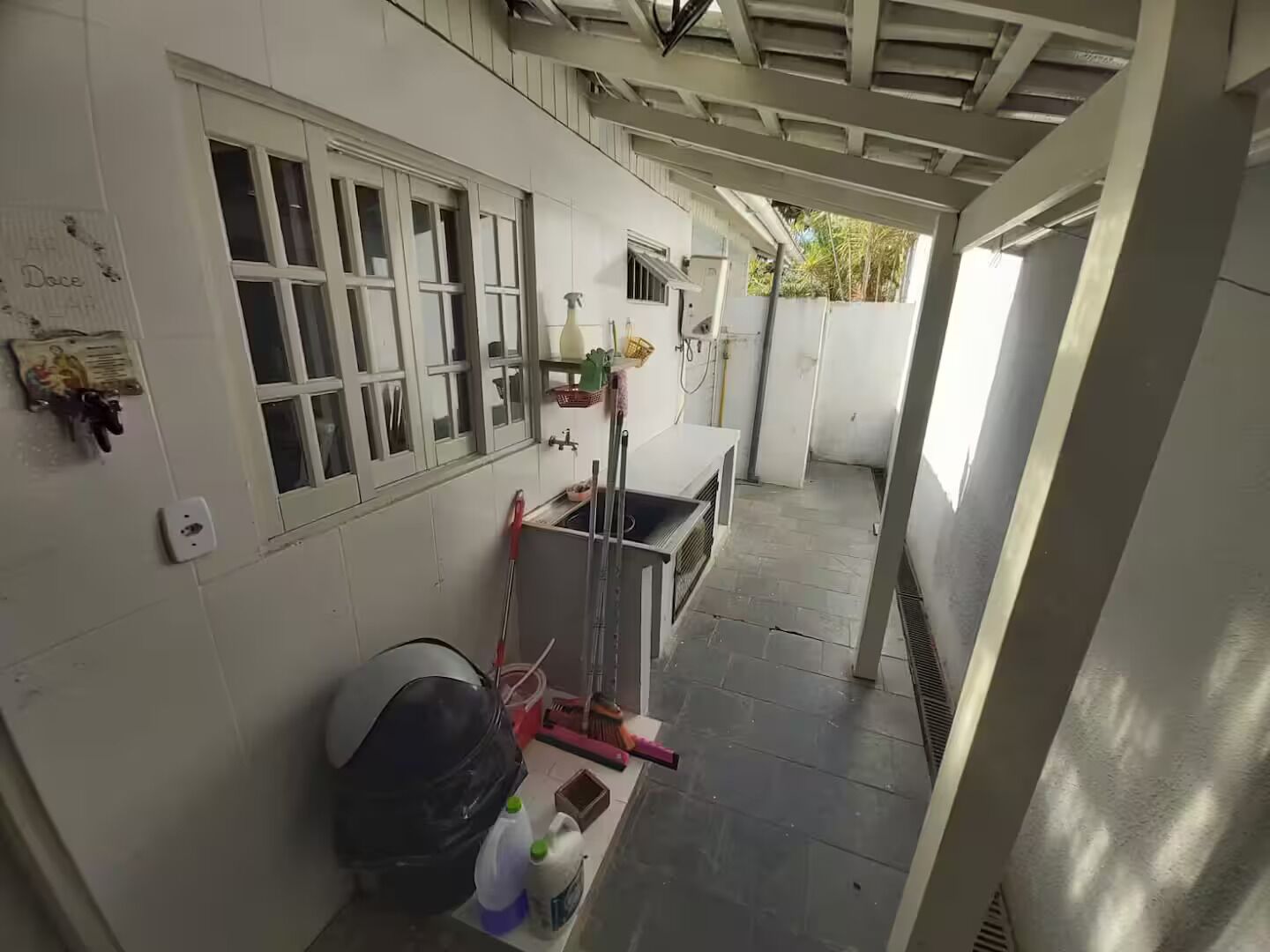 A narrow corridor with a tiled floor, a window with bars, a wall-mounted shelf with cleaning supplies, and a door leading to another room.