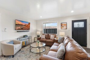 A living room with a beige sofa, a brown leather couch, a glass coffee table, and a large TV mounted on the wall.