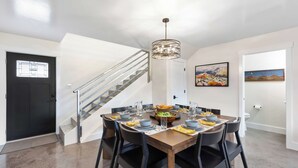 Un coin repas avec une table en bois dressée pour quatre personnes, entourée de chaises noires, d’un lustre moderne et d’un escalier avec des rampes en métal.