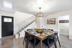 Comedor con mesa de madera dispuesta para cuatro, sillas negras, una lámpara moderna y una escalera con barandilla metálica.