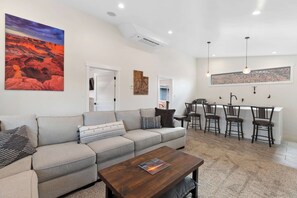 A modern living room with a sofa, coffee table, and a painting of a landscape. There is a kitchen area with bar stools and a window above the counter.