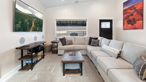 Un salon moderne comprenant un canapé d’angle, une table basse, un téléviseur posé sur un meuble, une fenêtre donnant sur un paysage montagneux et une porte à cadre noir.