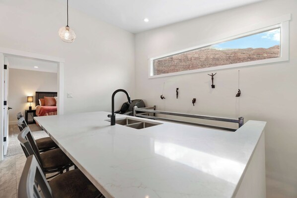 A modern kitchen with a large island, a dining area with chairs, a pendant light, a window with a view, and a bed in the adjacent room.