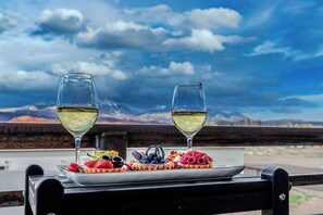 A table with two glasses of white wine and a plate of assorted fruits and pastries, set against a scenic mountain backdrop.