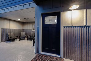 A building entrance with a dark blue door, a small window above it, and a light fixture above the door. There are two chairs outside the entrance.