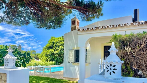 A white building with a pool, surrounded by greenery and a clear sky.