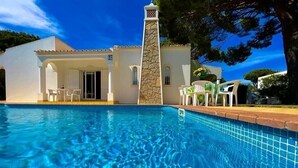 A swimming pool with clear blue water, a white building with a stone tower, and outdoor seating with chairs.