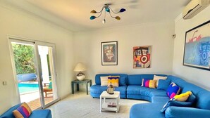 A living room with a blue sofa, a coffee table, and a lamp. There is a painting on the wall and a window with a view of a pool.
