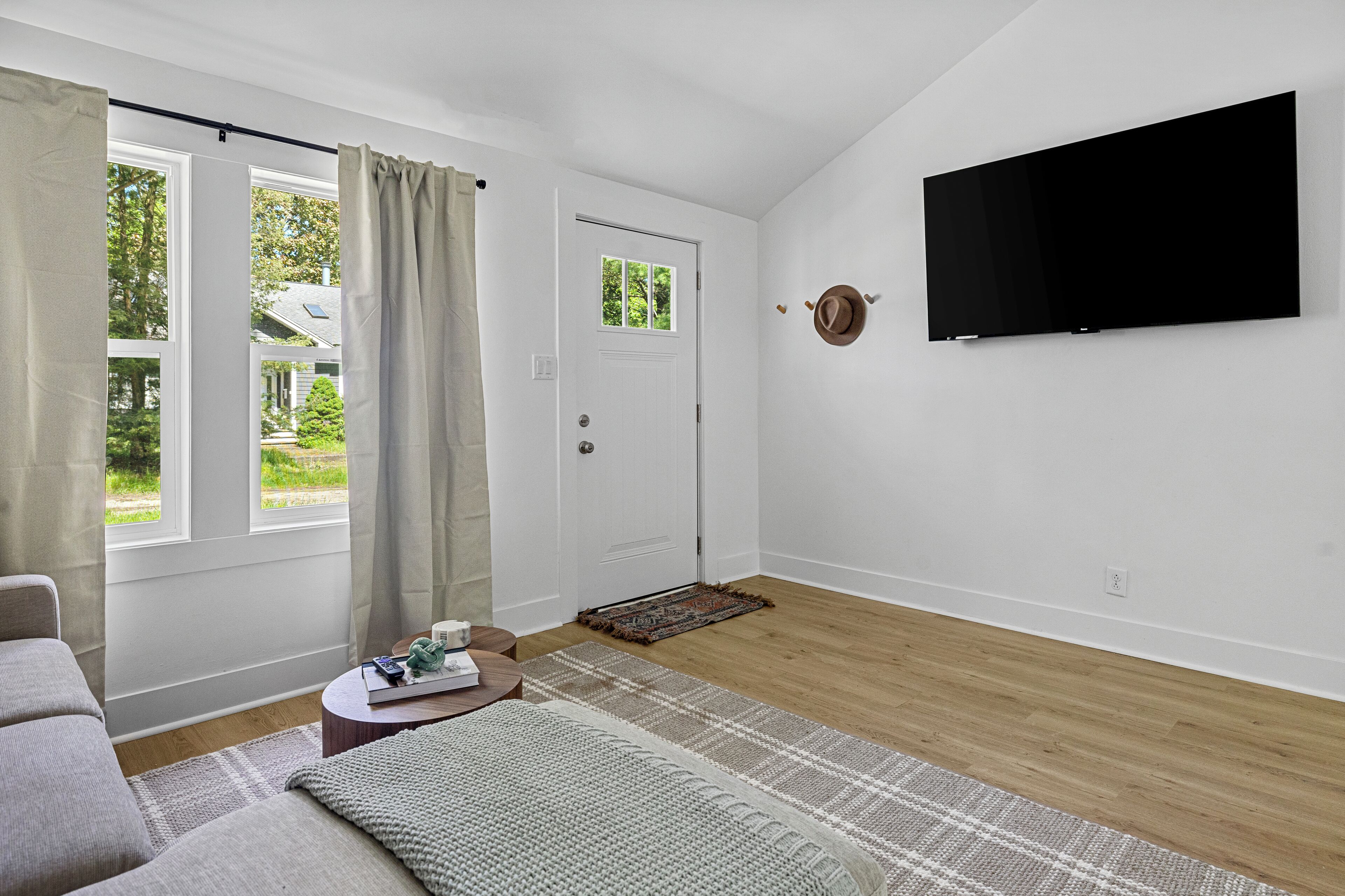 Een moderne woonkamer met een flatscreen televisie aan de muur, een bank, een salontafel en een groot raam met gordijnen.