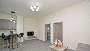 A modern living room with a grey sofa, a wooden coffee table with flowers, and a kitchen area with grey cabinets and a TV.