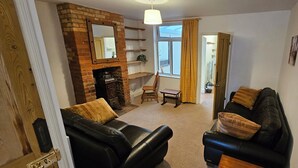 A living room with a brick fireplace, two black leather sofas, a wooden chair, a small table, and a large window with curtains. - Beautiful Spacious 5 Bed House (Bury St Edmunds)