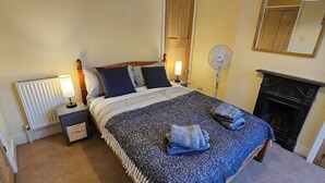 A bedroom with a bed, bedside lamps, a standing fan, a fireplace, and a mirror on the wall. - Beautiful Spacious 5 Bed House (Bury St Edmunds)