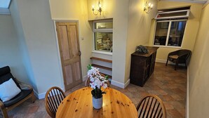 A dining area with a wooden table, chairs, and a vase of flowers. There is a door, a window, and a chair with a cushion. - Beautiful Spacious 5 Bed House (Bury St Edmunds)