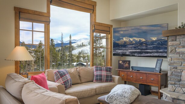 Un salon chaleureux avec un canapé beige, un banc en bois et une cheminée en pierre. De grandes fenêtres offrent une vue sur des montagnes enneigées.