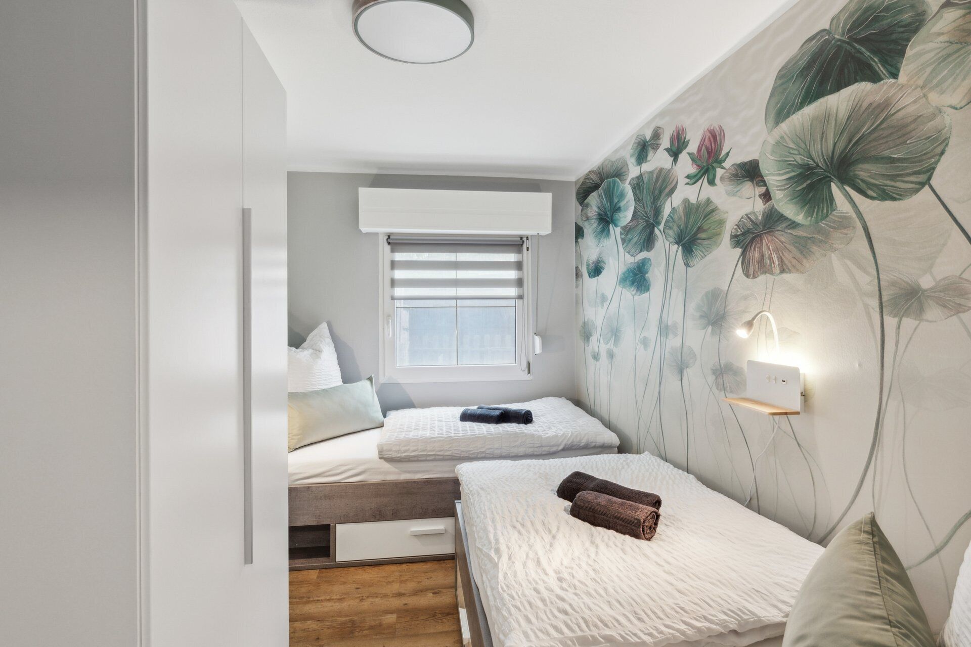 A bedroom with two beds, a window with blinds, a wall-mounted light, and a floral wallpaper.