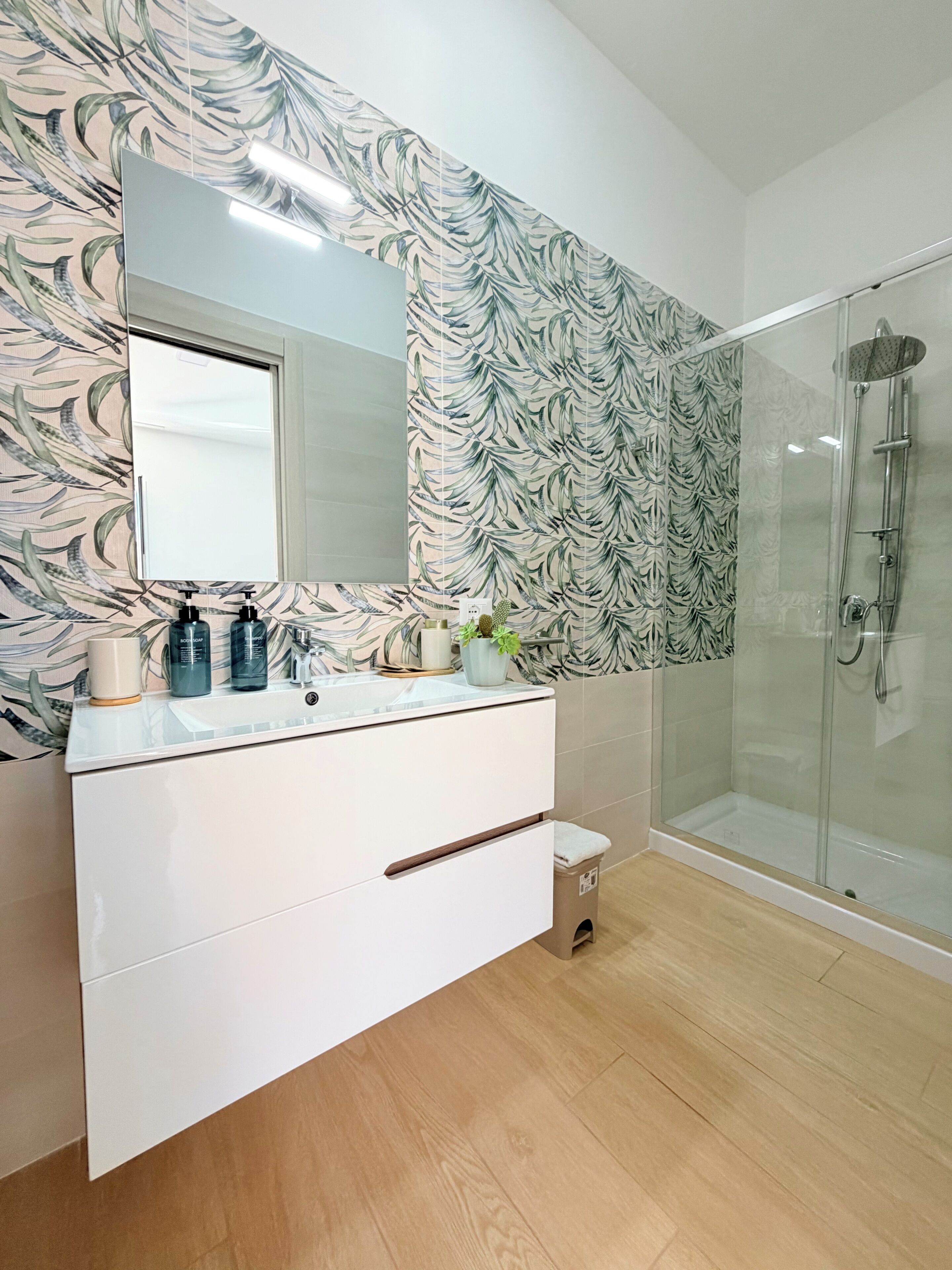 A modern bathroom with a large mirror, a sink with toiletries, a shower, and a patterned wall.
