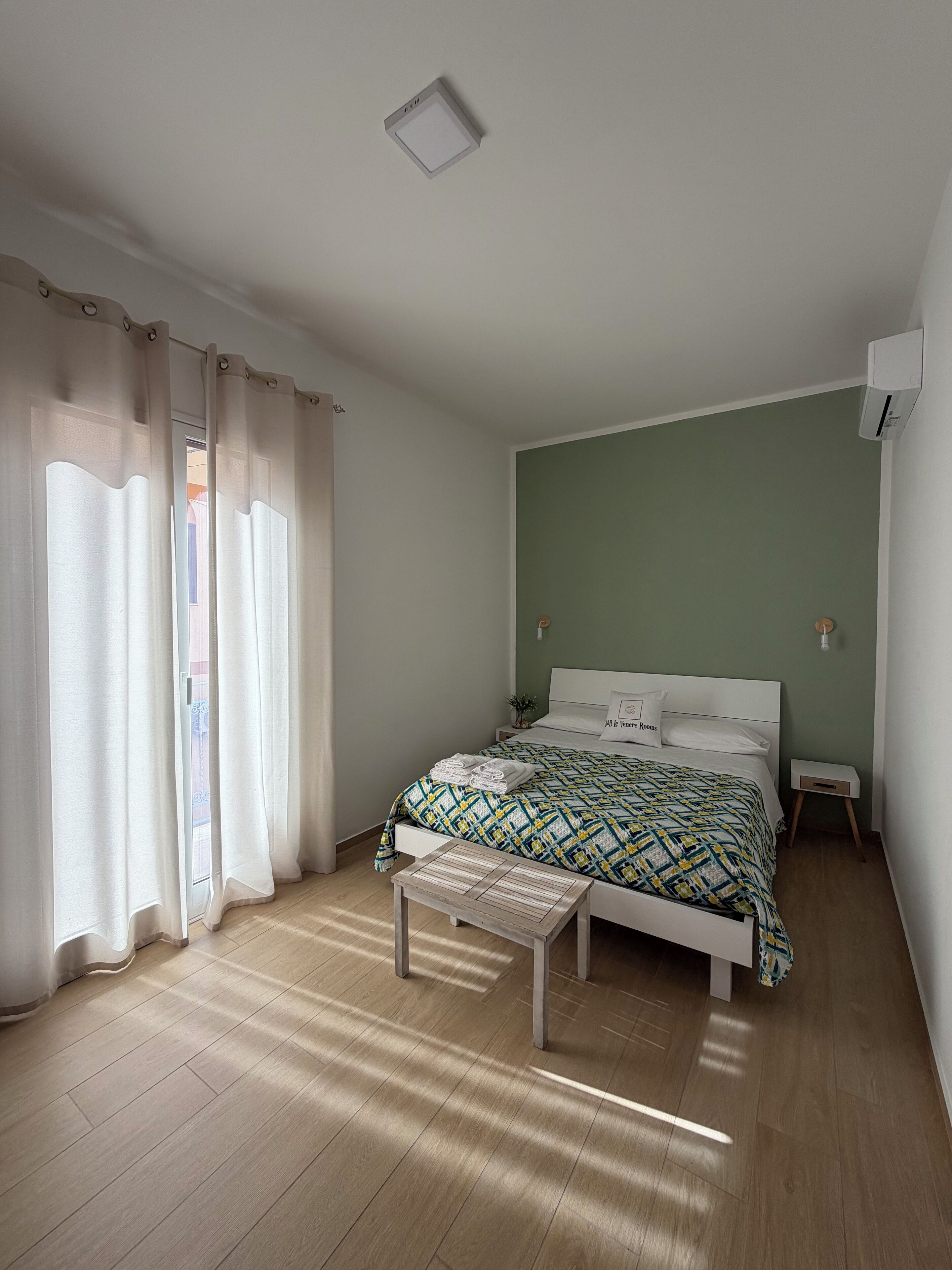 Een slaapkamer met een bed, nachtkastje, een bankje en een raam met gordijnen.