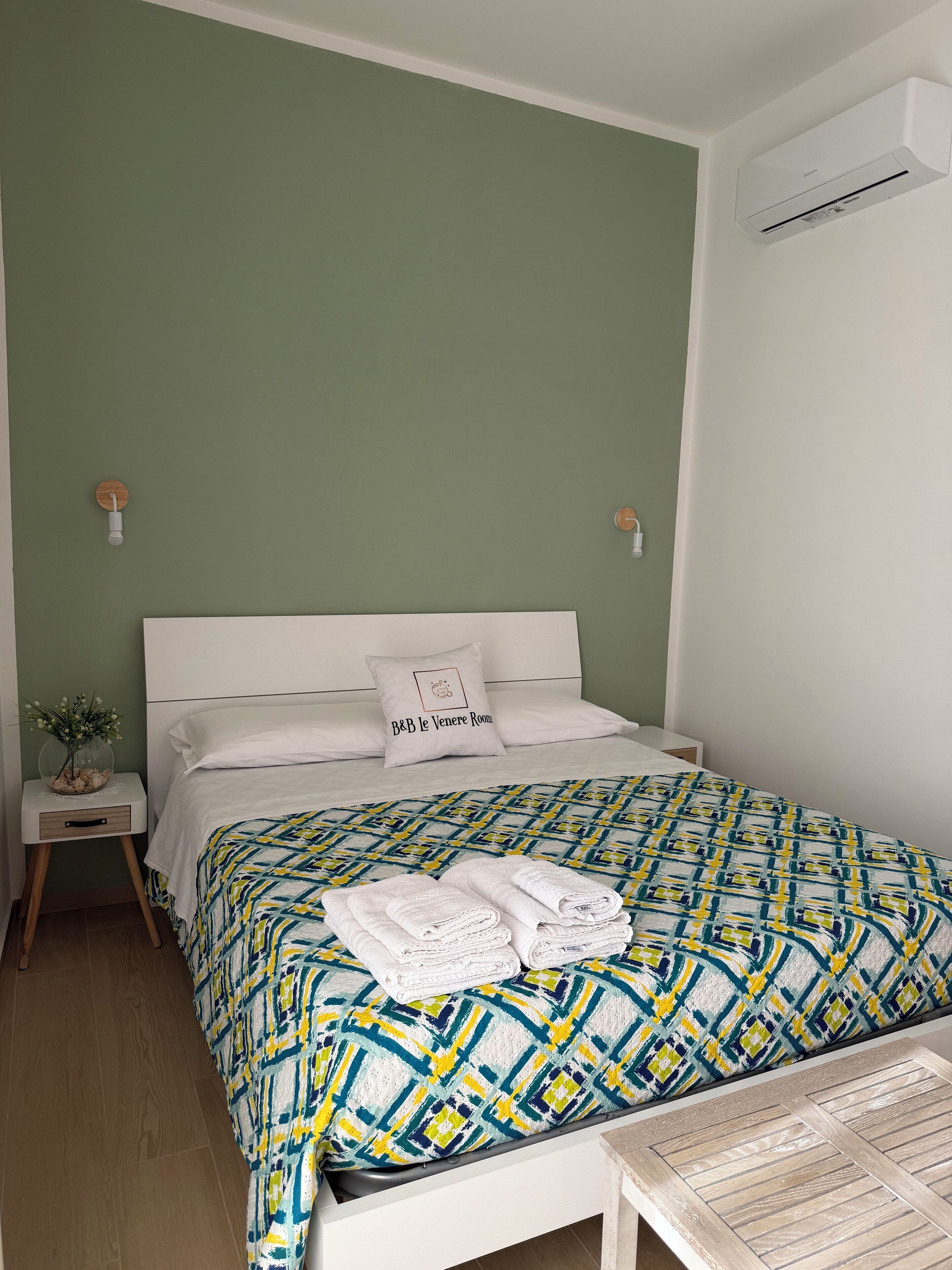 A neatly made bed with a patterned bedspread, a pillow with text, a bedside table with a vase, and an air conditioning unit on the wall.