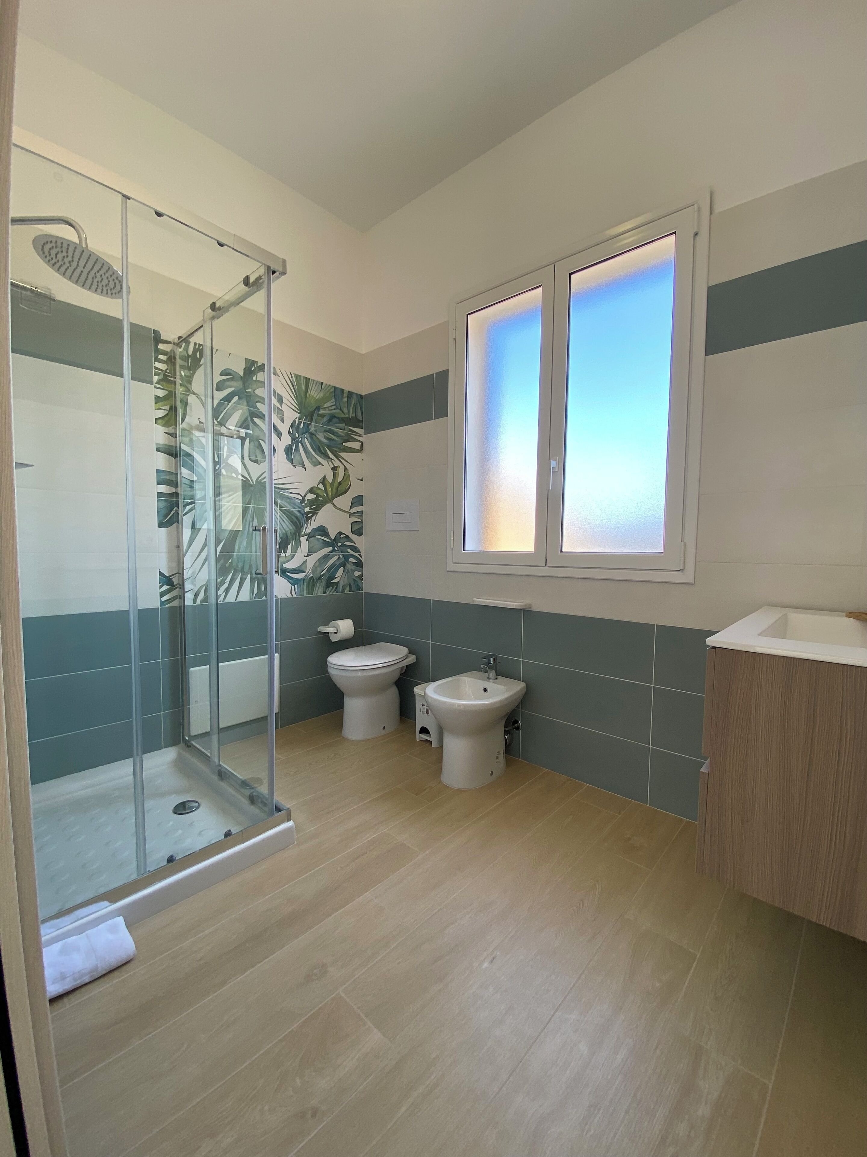A bathroom with a glass shower enclosure, two toilets, a bidet, and a window with frosted glass.