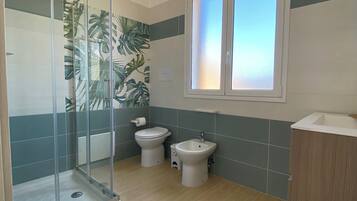 A bathroom with a glass shower enclosure, two toilets, a bidet, and a window with frosted glass.