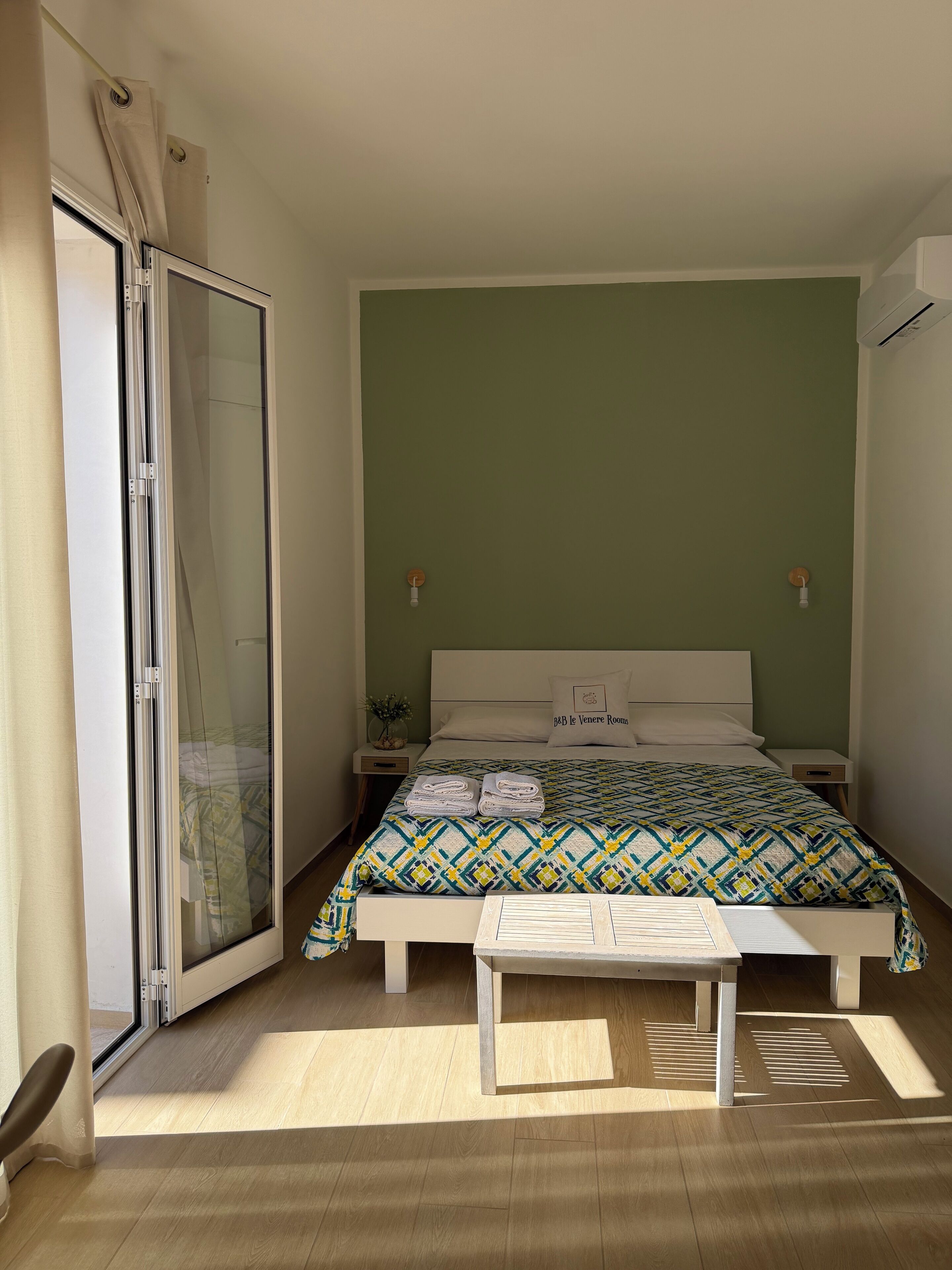 A bedroom with a bed, a bedside table, a nightstand, a sliding glass door, and a wall-mounted light fixture.