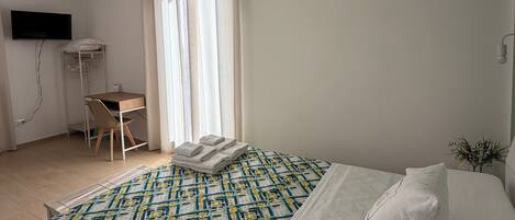 Quarto de hotel com cama, escrivaninha, cadeira, televisão e janela com cortinas.