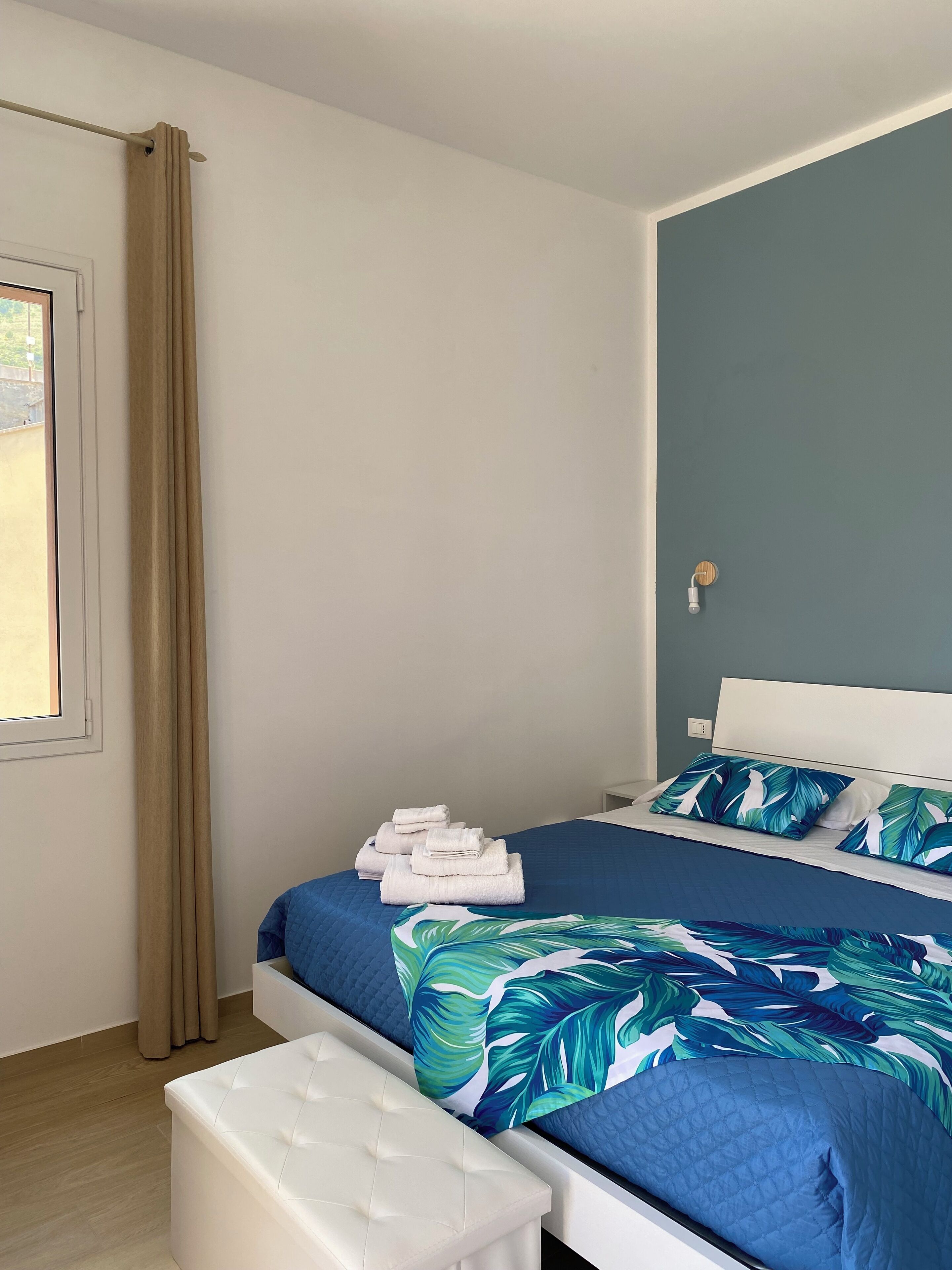A neatly made bed with a blue and green leaf-patterned bedspread, a white headboard, and a small stack of folded white towels on the foot of the bed.