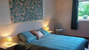 Ein Schlafzimmer mit einem Bett, Nachttischen, einem Sessel, einem Fenster mit Vorhängen und einem Wandbehang mit Blumenmuster.