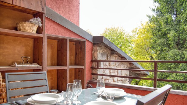Una mesa de comedor dispuesta con platos, vasos y cubiertos, con vistas a árboles a través de la barandilla de un balcón.