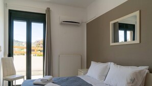 A hotel room with a bed, a chair, a window with curtains, an air conditioning unit, a radiator, and a mirror on the wall. - Three Waves Maisonettes Elena (Agios Nikolaos)