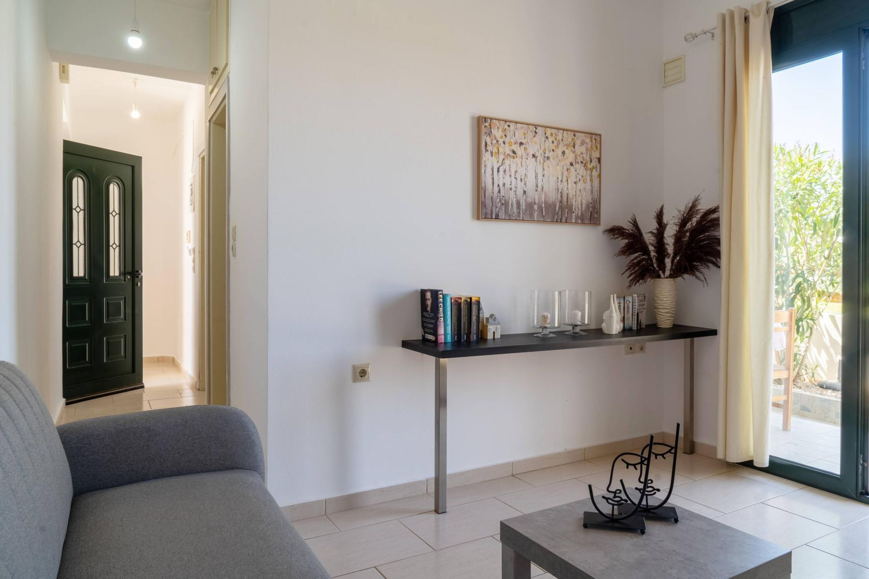 A living room with a sofa, a coffee table, a console table with books and decorative items, a painting on the wall, and a view of the outdoors through a door.