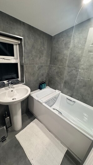 A modern bathroom with a white sink, a glass-enclosed bathtub, and grey tiled walls. - PS5+Games+Parking|Sleeps 6, 60"TV + Netflix, ideal for Business/Relocator stays (London)