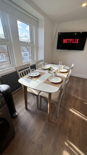 Uma área de jantar com uma mesa posta para quatro pessoas, uma televisão fixada na parede e uma janela com vista para edifícios.