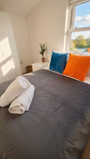 A neatly made bed with a grey bedspread, two rolled white towels, and two pillows in blue and orange. - PS5+Games+Parking|Sleeps 6, 60"TV + Netflix, ideal for Business/Relocator stays (London)