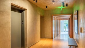 A modern hallway with a wooden floor, a reception desk, and a glass door marked with an elevator sign.