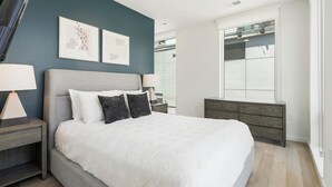 A modern bedroom with a large bed, two bedside tables, a dresser, and a window with blinds.