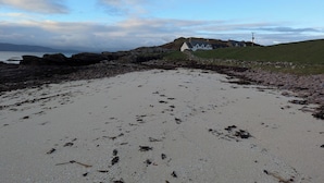 Ein sandiger Strand mit vereinzelten Tangstrudeln, ein weißes Gebäude mit grüner Dachbegrünung und eine felsige Küstenlinie.