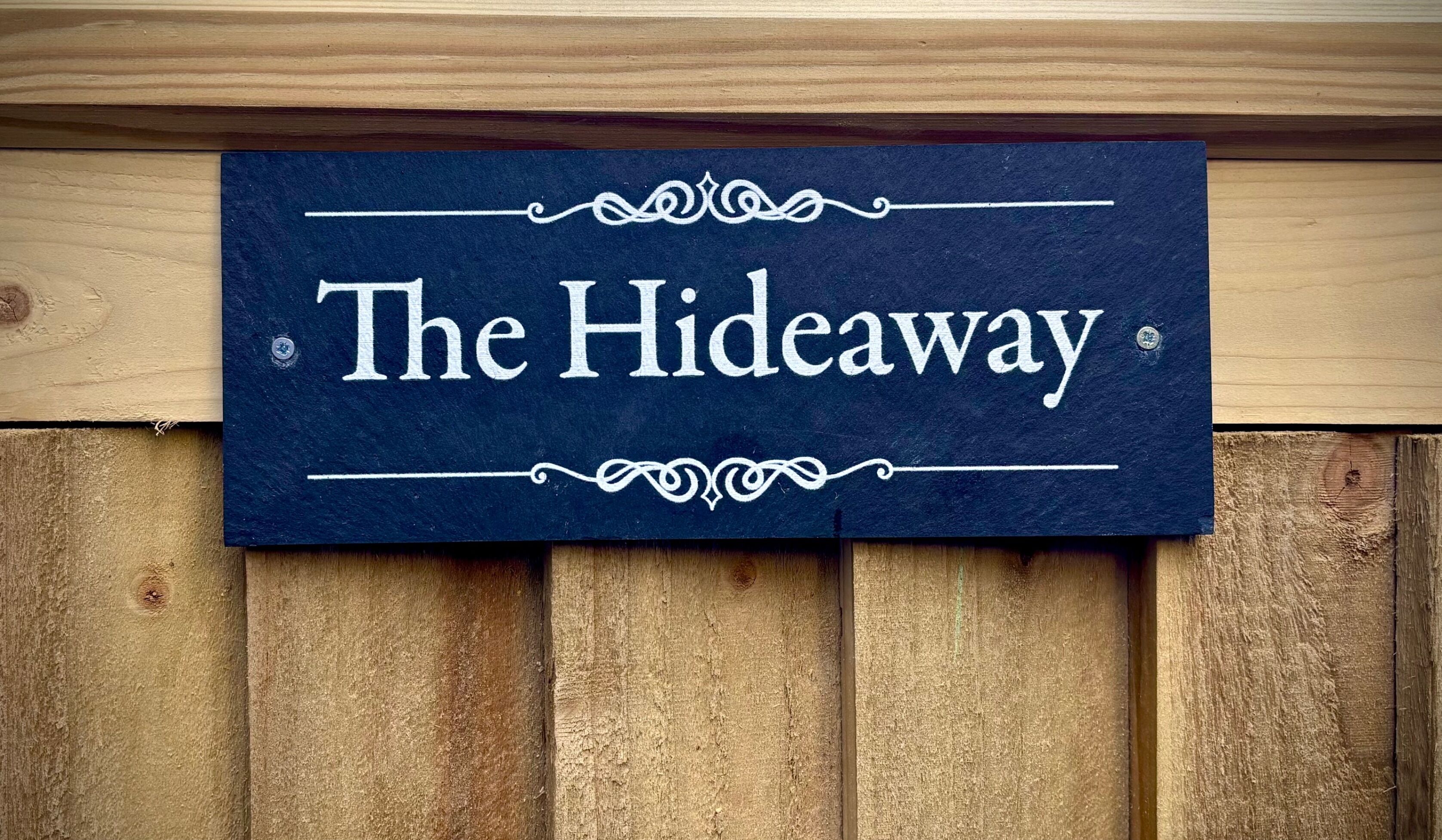 Un letrero azul oscuro con la palabra "The Hideaway" en blanco, rodeado de adornos en espiral, fijado a una pared de madera.