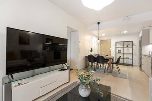 A modern living room with a flat-screen TV, a glass coffee table, and a dining area with a wooden table and chairs.