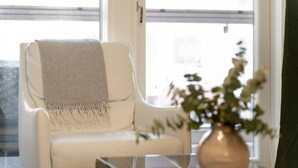 A white armchair with a grey throw blanket, a clear glass coffee table, and a vase with flowers. - Superior Queen Room in Tammela Soccer Stadium! (Tampere)